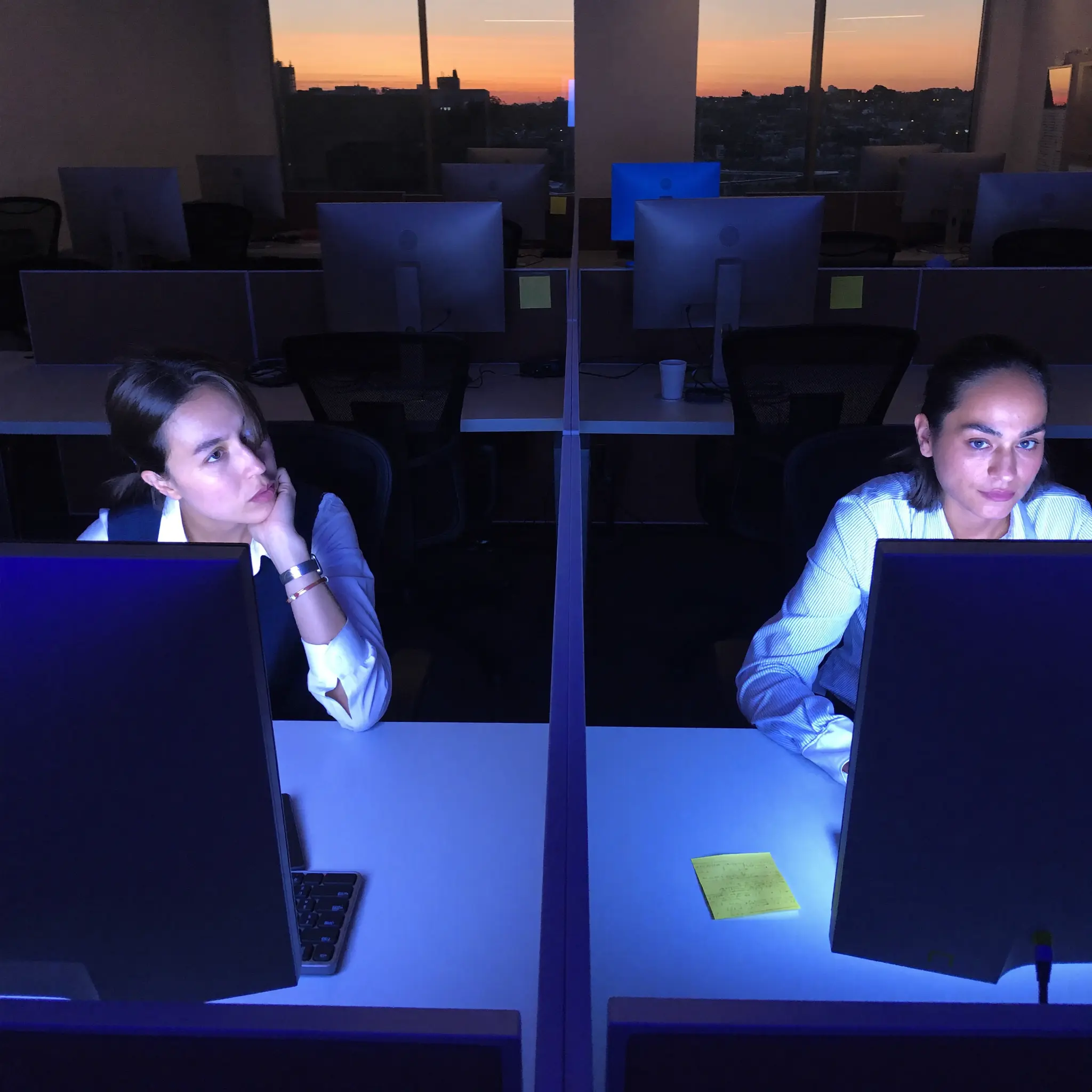Two coworkers at adjacent desks in a strained after-hours office moment.